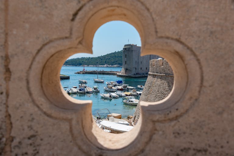 Historic Dubrovnik harbor viewed through a decorative stone opening, showcasing boats and the ancient fortress.